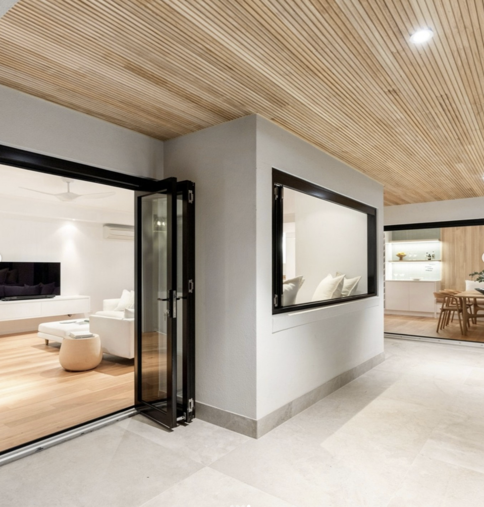 Open-plan coastal home with large-format stone-look porcelain floor tiles running from the living area out to the alfresco, under a timber batten ceiling